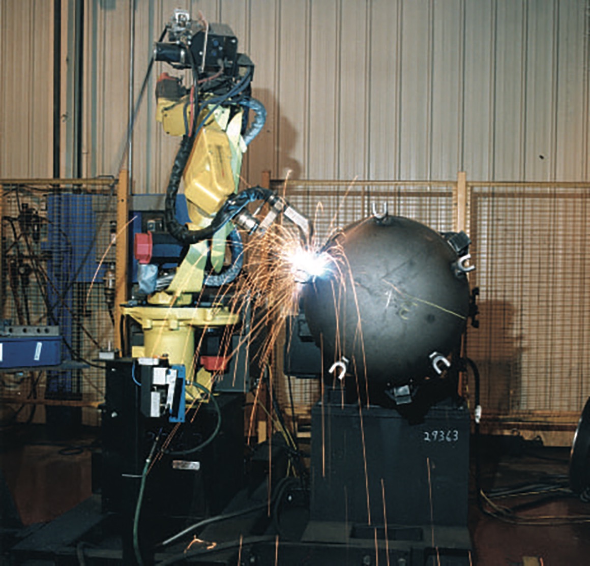 Industrial robotic arm performing welding on a spherical component, showcasing the technology behind quick opening closures in manufacturing. Sparks fly as the automated process ensures precision and efficiency in production.
