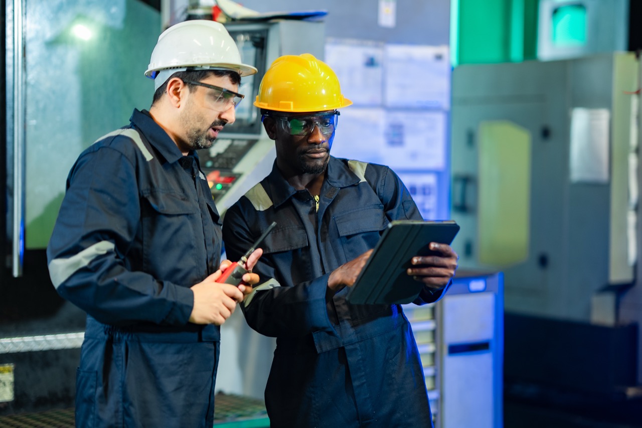 Two industrial workers wearing safety helmets and protective eyewear are collaborating in a manufacturing setting. One worker is holding a tablet while the other is using a handheld tool, demonstrating teamwork and communication in a technical environment.
