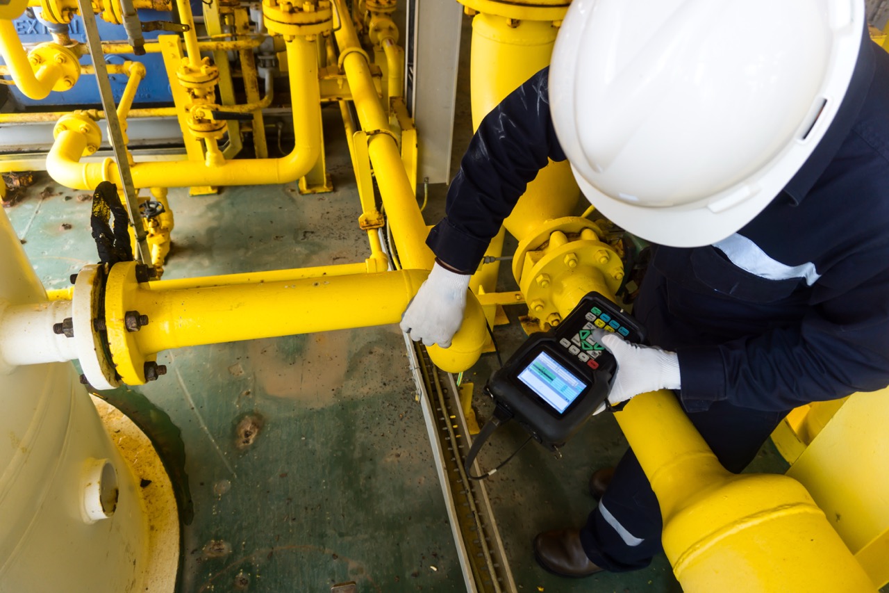 A technician in a white hard hat and gloves inspecting yellow industrial pipes using a handheld digital device in a factory setting.