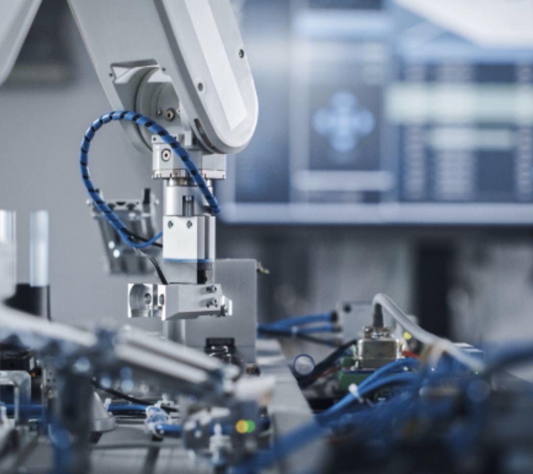 Close-up view of a robotic arm in a high-tech manufacturing environment, showcasing intricate mechanical components and blue wiring, with a blurred background of digital screens displaying data and controls.