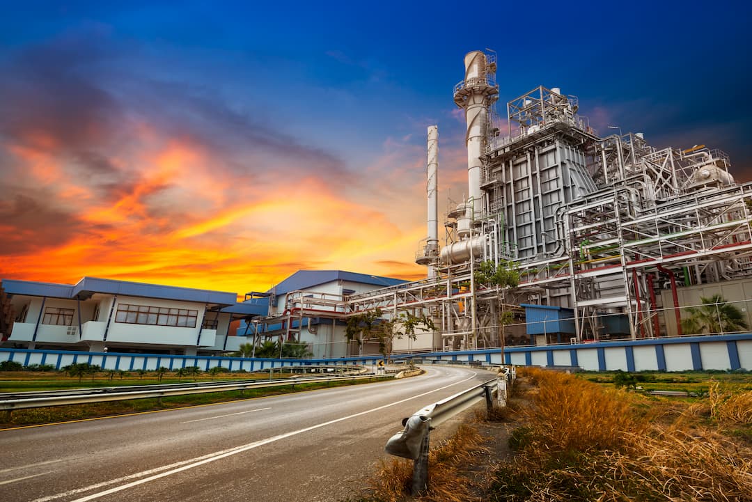 Industrial chemical processing facility at sunset, showcasing large equipment and pipes, surrounded by a winding road and greenery.