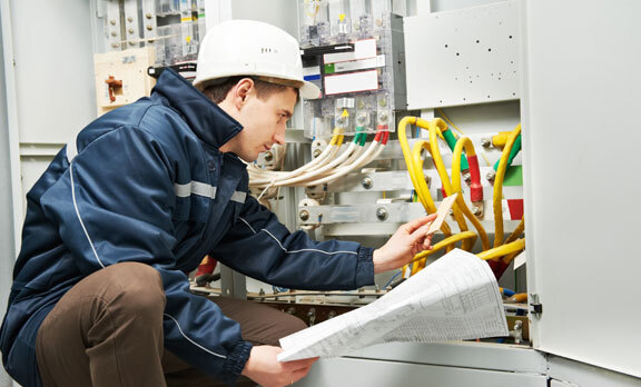 Electrician inspecting wiring in a control panel while reviewing technical documentation, wearing a hard hat and safety gear.