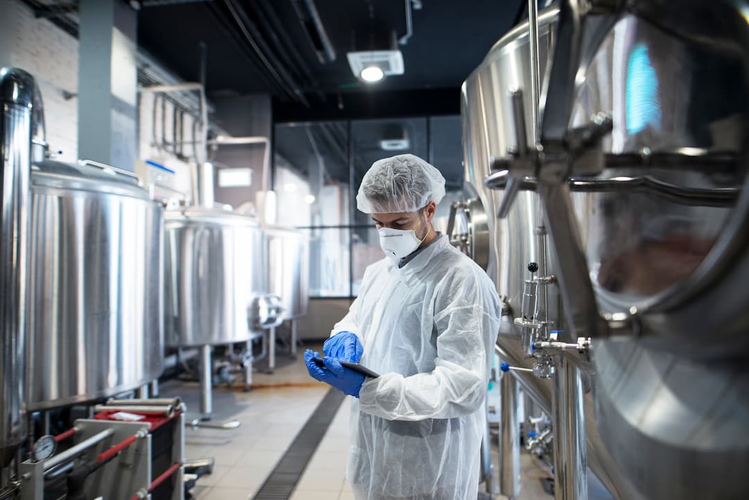 Worker in protective gear using a tablet in a modern brewery, surrounded by stainless steel fermentation tanks. The image highlights safety protocols and technology in beverage production.