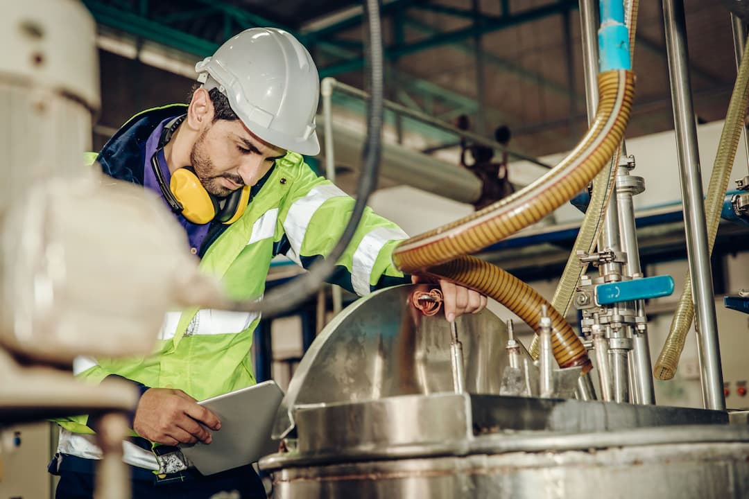 A worker in a safety helmet and high-visibility jacket inspects equipment in an industrial setting, using a tablet to monitor processes in a manufacturing facility.