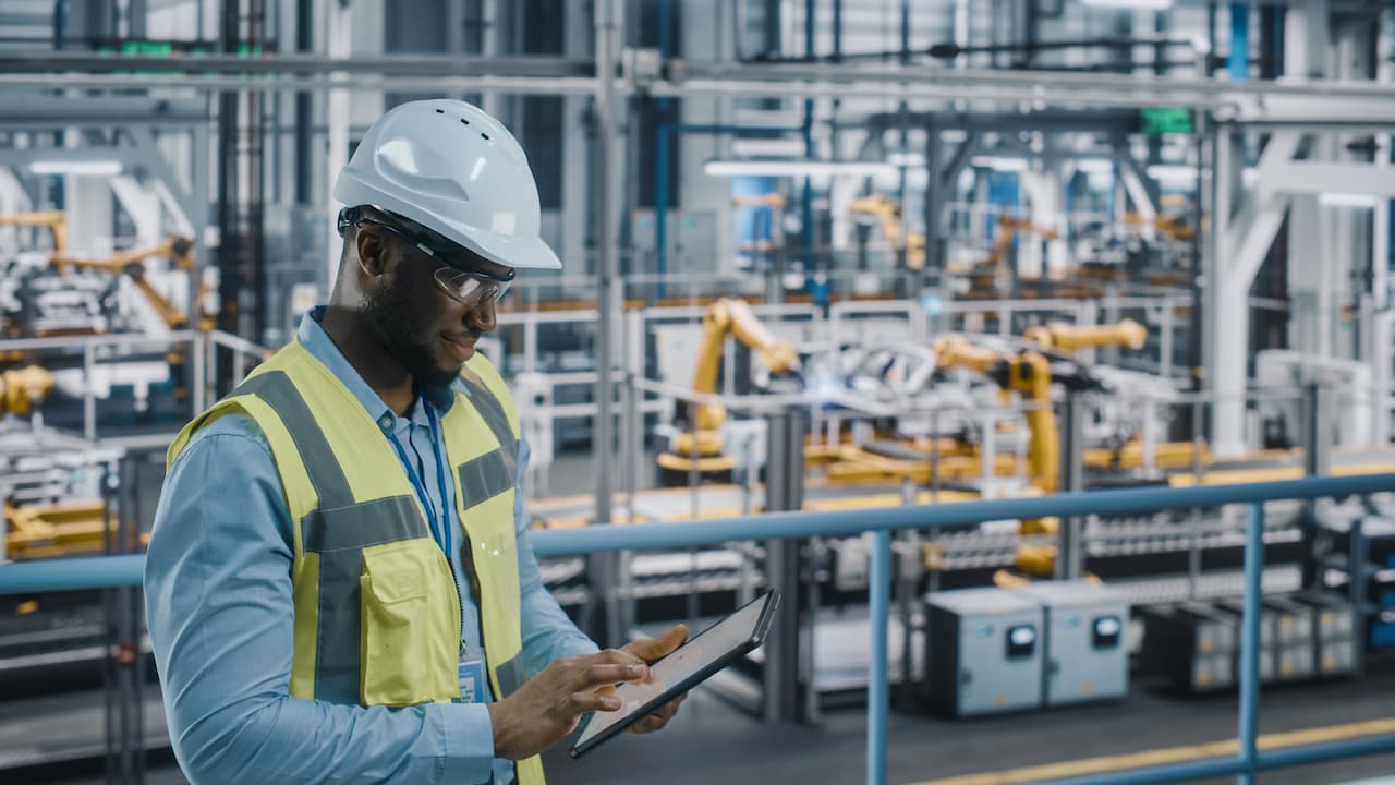 A construction worker wearing a hard hat and safety goggles uses a tablet to monitor operations in a modern manufacturing facility with robotic machinery in the background.