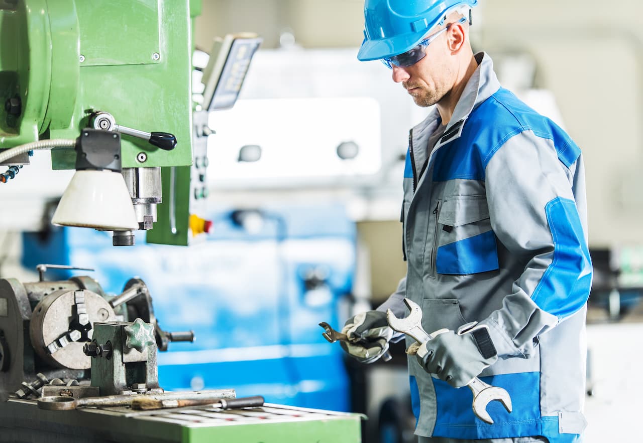 Worker in safety gear using wrenches in a manufacturing facility, surrounded by machinery and tools, emphasizing industrial craftsmanship and safety practices.