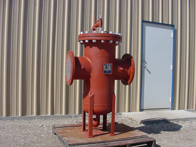 Oil basket strainer displayed outdoors, featuring a red cylindrical body with multiple inlet and outlet ports, designed for filtering oil in industrial applications. The strainer is mounted on a wooden platform against a backdrop of a metal wall and a door.