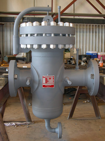 Gray meter protection basket strainer mounted in a workshop, showcasing its inlet and outlet connections, filter basket chamber, and pressure gauge, designed for efficient fluid filtration in industrial applications.