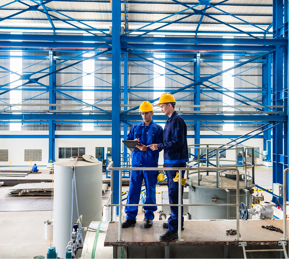 Two construction workers in blue overalls and yellow hard hats review plans on a tablet, standing on a platform in a large industrial warehouse with blue steel beams and machinery in the background.