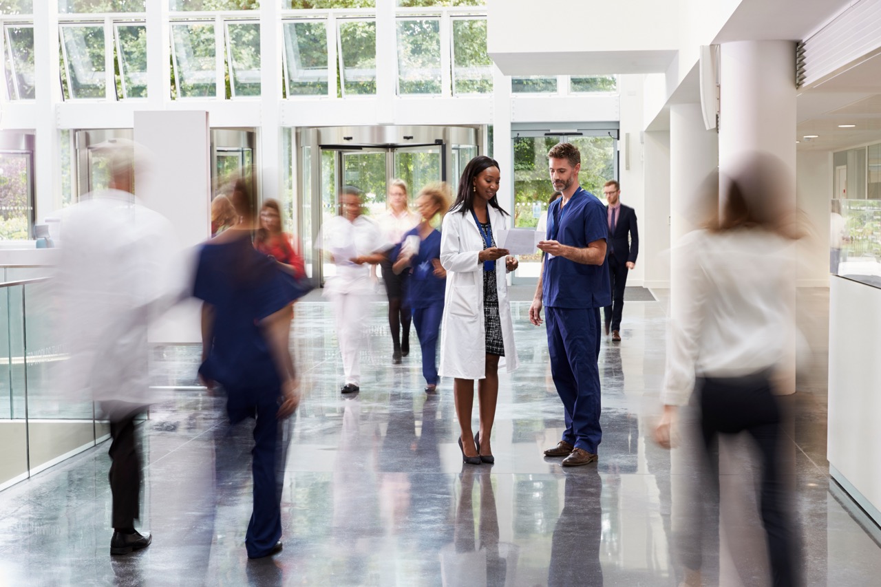 Healthcare professionals in a busy hospital corridor, with a female doctor in a white coat discussing patient information with a male nurse in scrubs, while other medical staff and visitors move around in the background.