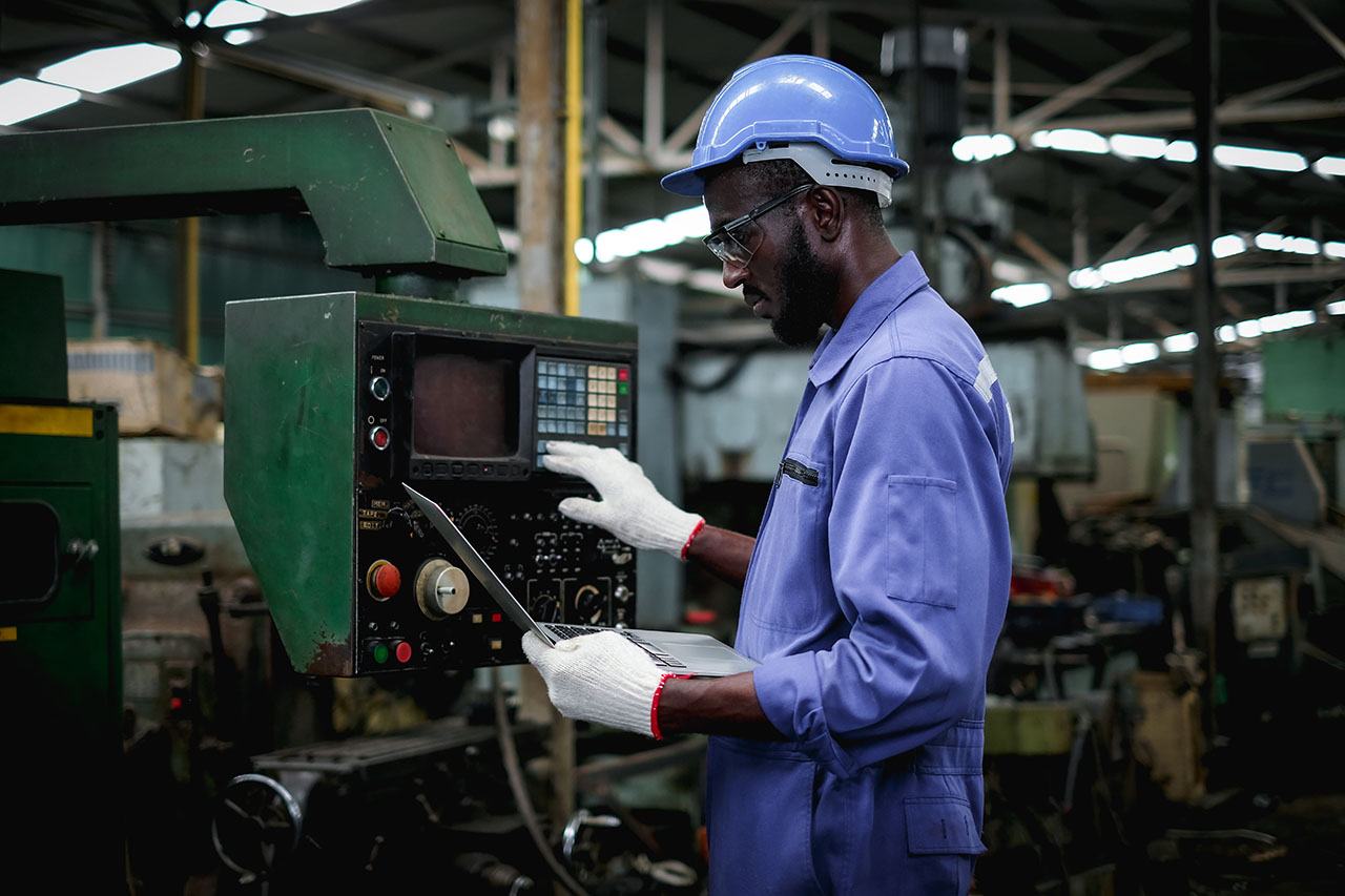 A factory worker in a blue jumpsuit and safety gear operates a machine control panel while holding a laptop, showcasing modern manufacturing practices and safety protocols in an industrial setting.