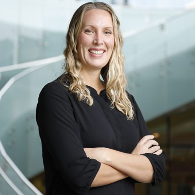 Professional woman smiling with arms crossed, standing in a modern office environment with glass walls, representing confidence and approachability.