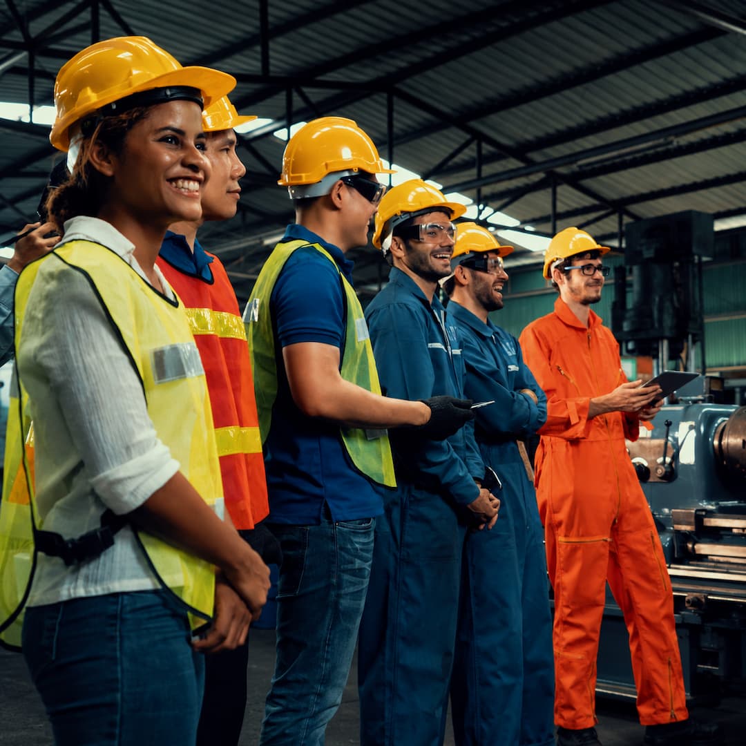 Diverse group of candidates in safety gear, including hard hats and reflective vests, smiling and engaged in a manufacturing environment, showcasing teamwork and readiness for industrial roles.