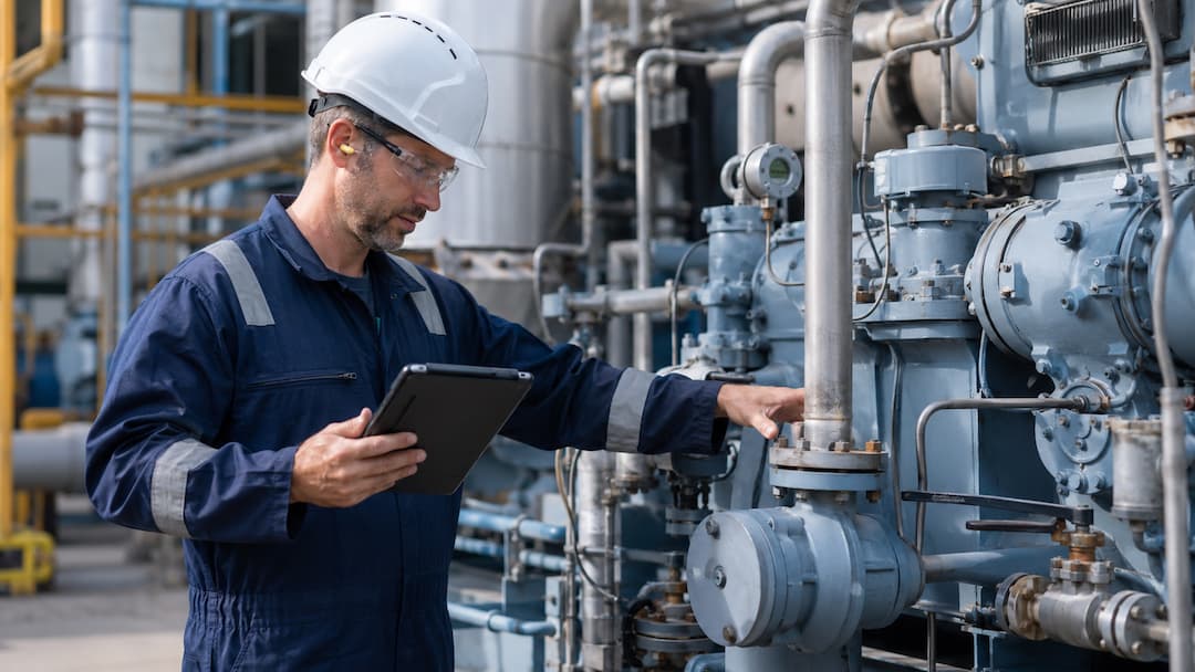 Engineer in safety gear using a tablet to monitor emissions solutions at an industrial facility, surrounded by machinery and equipment.