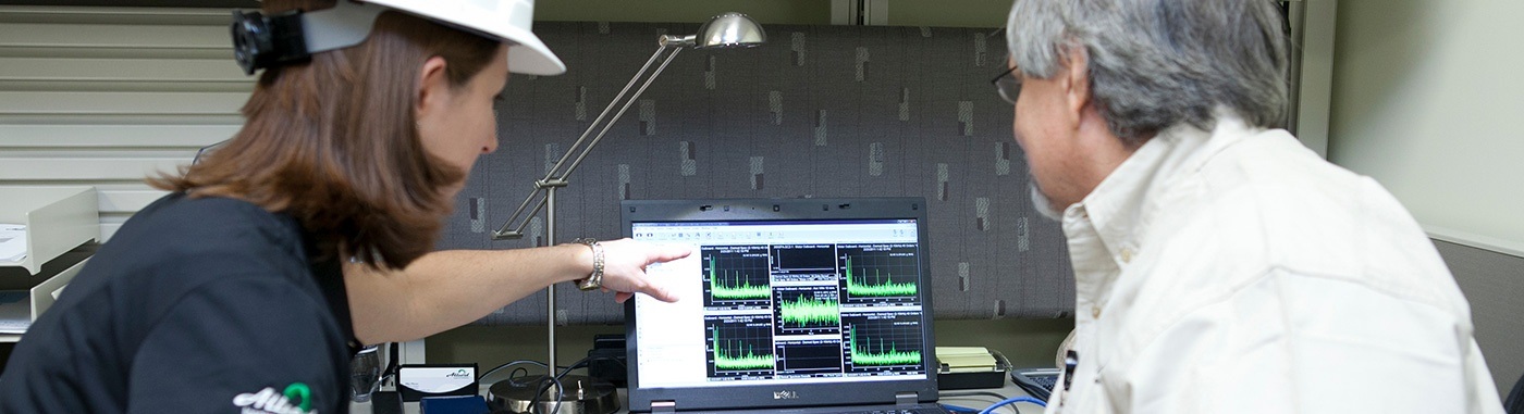 Two professionals analyzing data on a laptop screen, displaying various graphs and charts, in a modern office environment. The woman in a hard hat points at the screen while the man observes, highlighting collaboration in technical analysis.