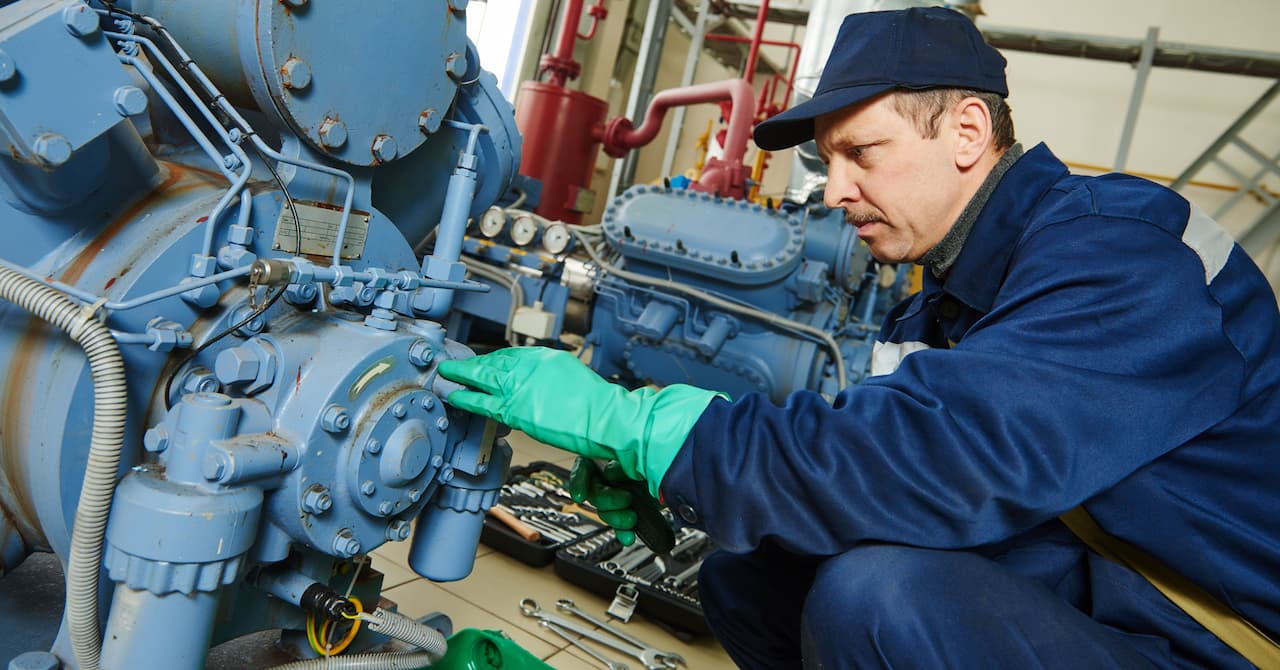 Maintenance technician inspecting industrial machinery with tools in a facility, wearing protective gloves and uniform, focused on equipment components.