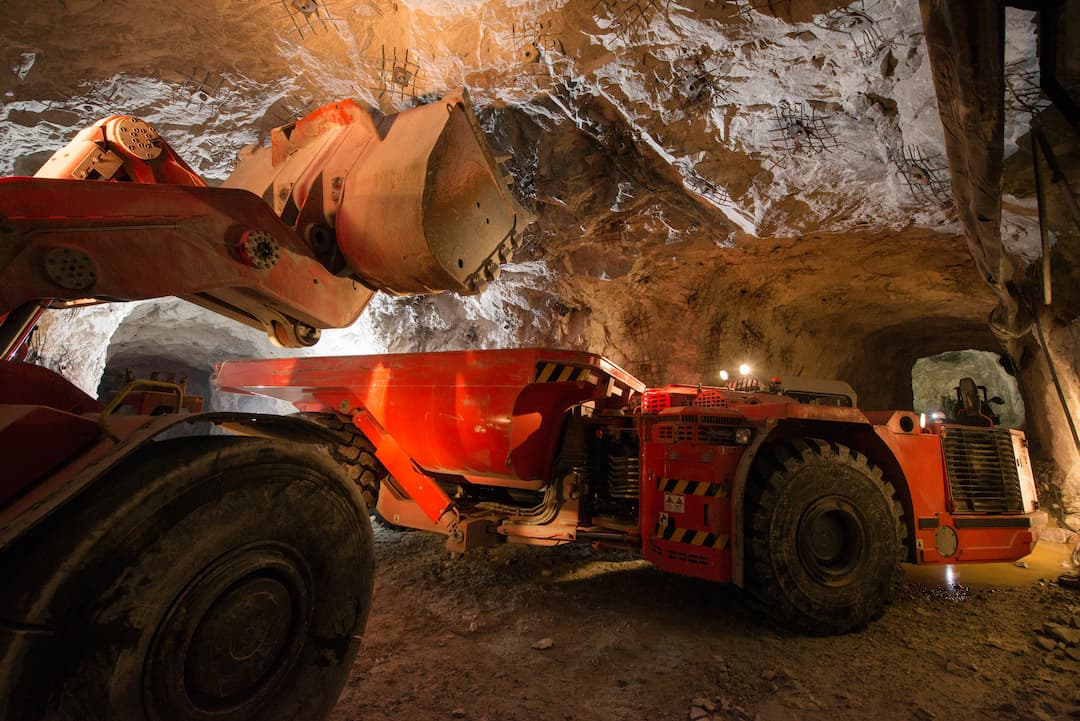 Heavy mining equipment operating in an underground mine, showcasing a red loader and a mining truck in a well-lit cavernous environment, emphasizing the machinery used in mineral extraction and materials handling.