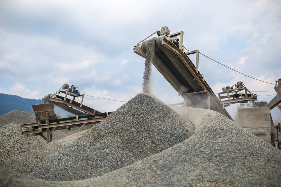 Conveyor belts transporting gravel at a mining site, with large piles of aggregate material in the foreground and a cloudy sky in the background, illustrating the mining and materials industry.