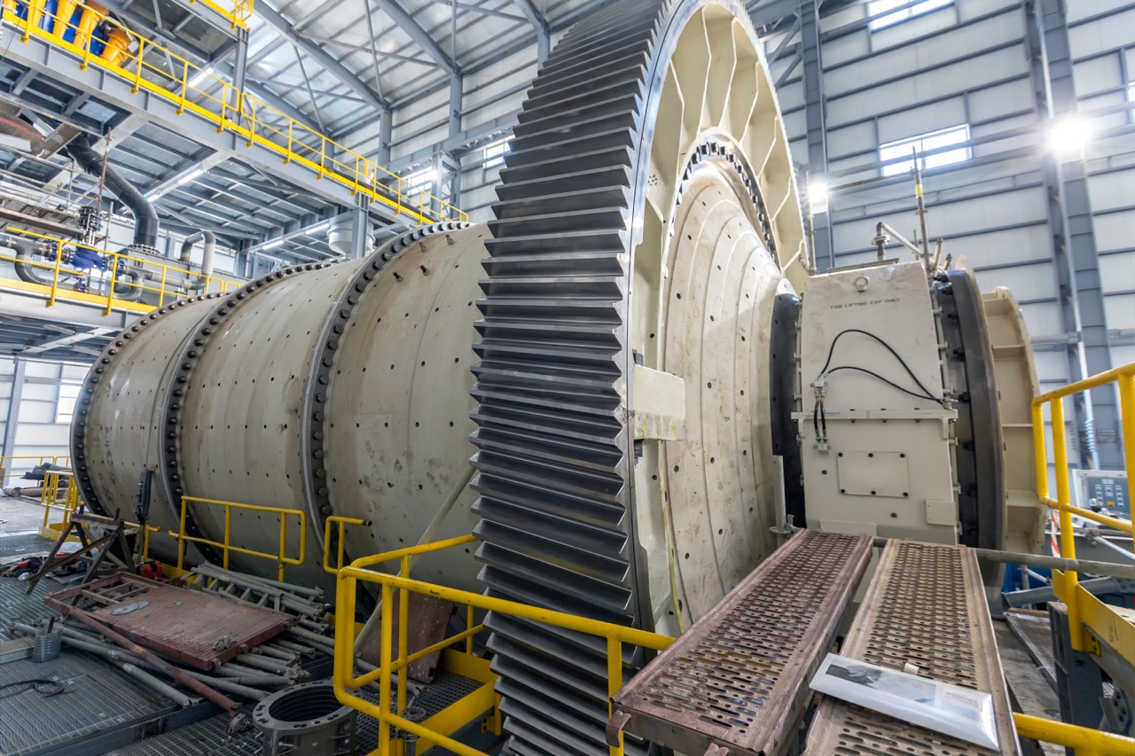 Industrial machinery with a large gear and cylindrical components inside a factory setting, showcasing the intricate design and engineering of heavy equipment used in manufacturing processes. Bright overhead lighting and structural elements are visible in the background.