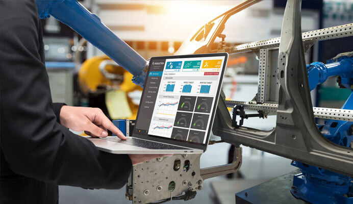 A person using a laptop to monitor manufacturing data in an industrial setting, with robotic arms and a vehicle chassis in the background, illustrating advanced automation and data analysis in modern production processes.