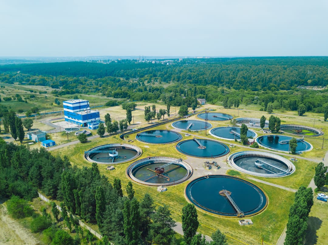 Aerial view of a modern wastewater treatment plant featuring multiple circular tanks, a blue processing facility, and surrounding green landscape, showcasing the infrastructure for water purification and environmental management.