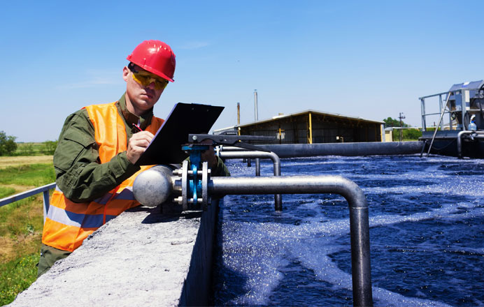 Worker in a red hard hat and safety vest inspecting water treatment facility, taking notes on a clipboard beside a tank with bubbling water.