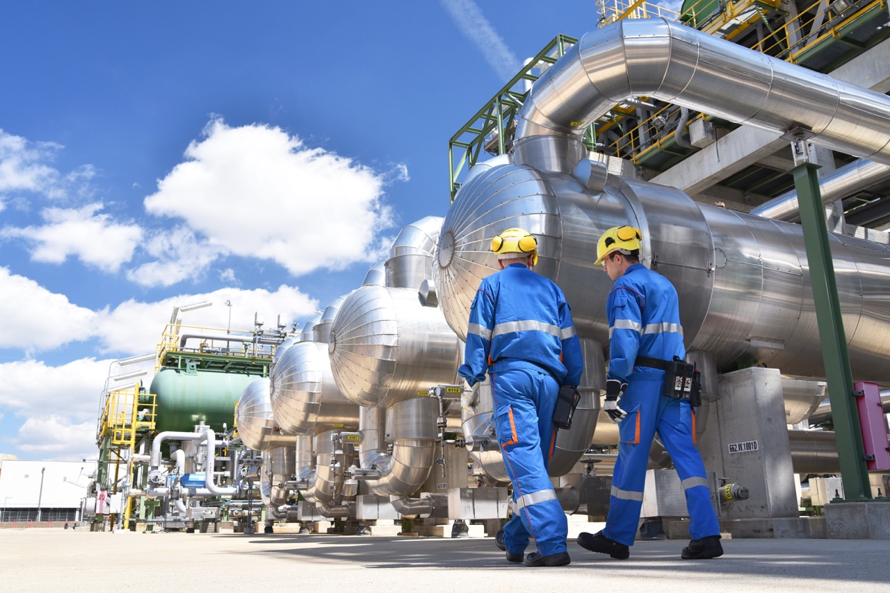 Two workers in blue uniforms and safety helmets inspect industrial piping and equipment at a gas processing facility under a clear blue sky.