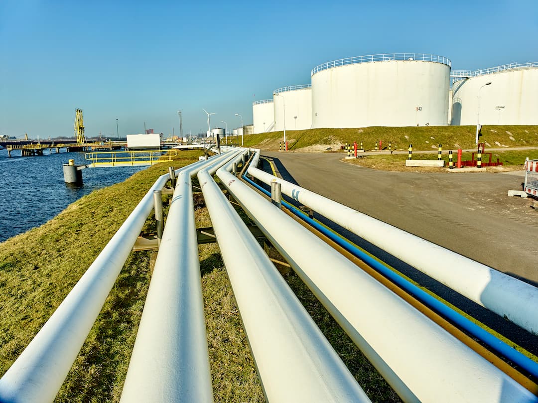 Industrial midstream infrastructure featuring a network of white pipelines leading to large storage tanks, with a clear blue sky and waterway in the background, illustrating the transportation and storage of oil and gas products.