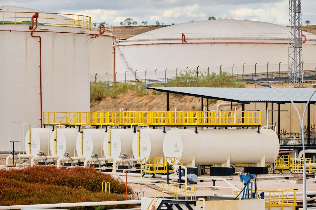 Storage tanks and processing equipment at a midstream oil facility, featuring large white tanks with yellow railings and pipelines, set against a backdrop of additional storage infrastructure.