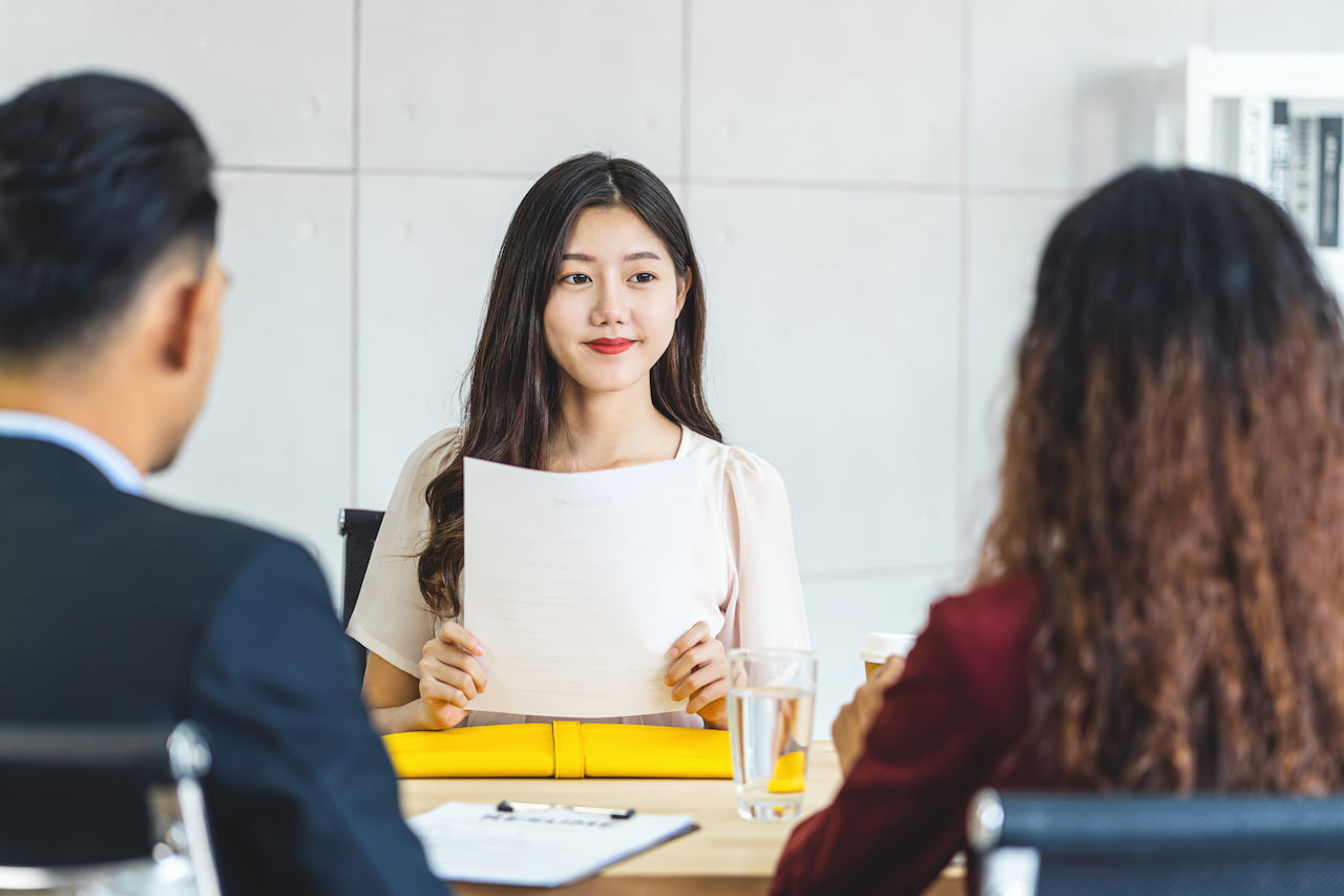 Woman sitting at a table in a job interview, holding a document, with two interviewers visible in the background. The setting features a modern office environment with a glass of water and a yellow folder on the table.