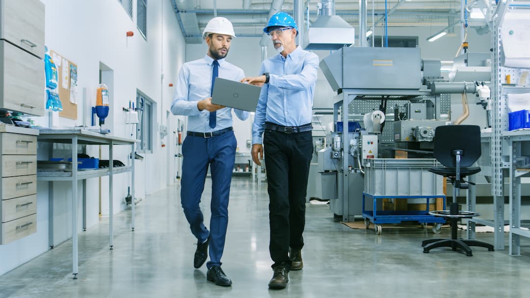Two professionals in hard hats walk through a modern industrial workspace, discussing a laptop. The setting features machinery and workstations, highlighting collaboration in a manufacturing environment.