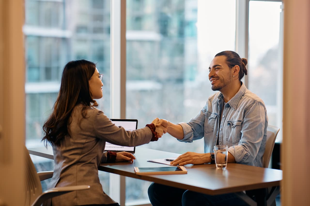Business professionals shaking hands during a meeting in a modern office, with laptops and notebooks on the table, symbolizing partnership and collaboration.