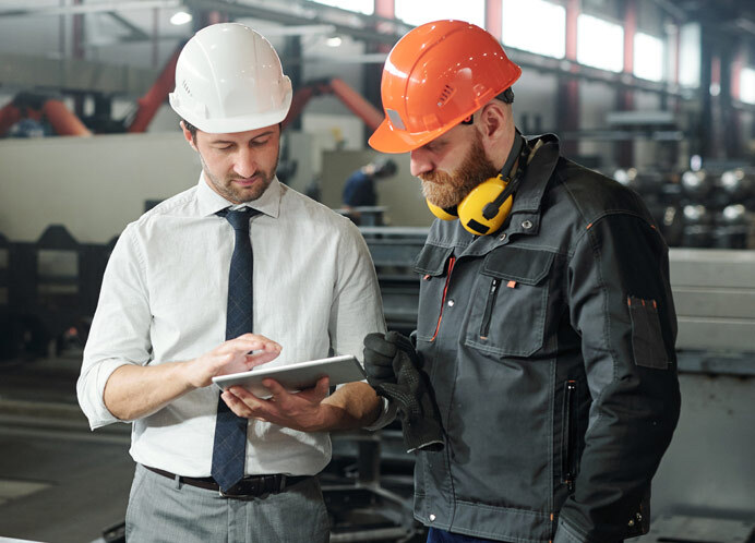 Two professionals in a manufacturing facility discuss staffing solutions while reviewing information on a tablet. One is dressed in a white hard hat and formal attire, while the other wears an orange hard hat and safety gear, highlighting the collaboration between management and skilled workers to address staffing challenges.
