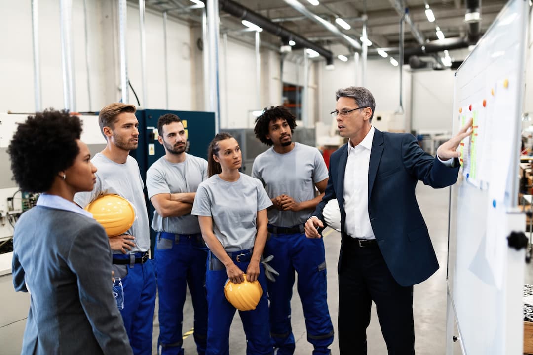 Group of factory workers and a manager discussing strategies to maximize throughput, with a focus on a whiteboard presentation in an industrial setting.