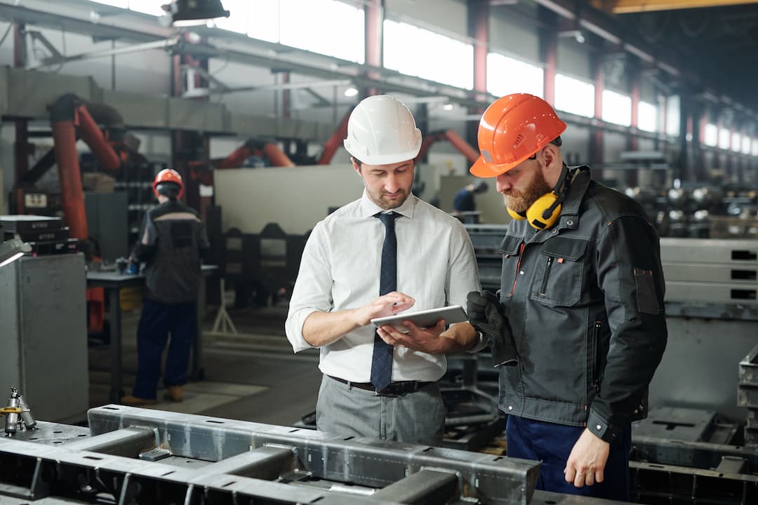Two professionals discuss strategies for maximizing throughput in a manufacturing facility, one wearing a white hard hat and the other in an orange hard hat with ear protection, while using a tablet to analyze data.