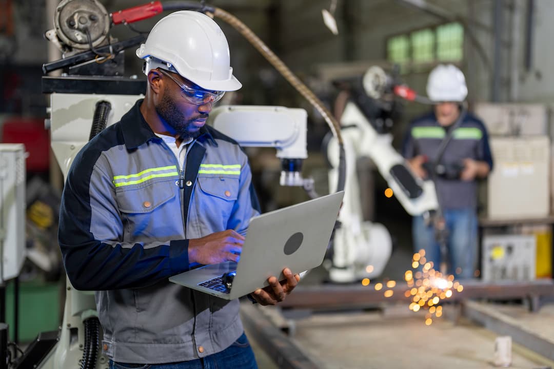 A worker in a hard hat and safety goggles operates a laptop in a manufacturing facility, with robotic arms in the background. The scene emphasizes the integration of technology in maximizing throughput in industrial settings.