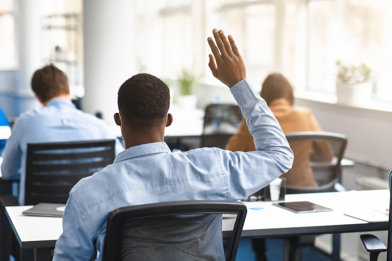 A student raising their hand in a modern classroom setting, with other students seated at desks, engaged in learning. The room features large windows, natural light, and a focus on participation and interaction in an educational environment.