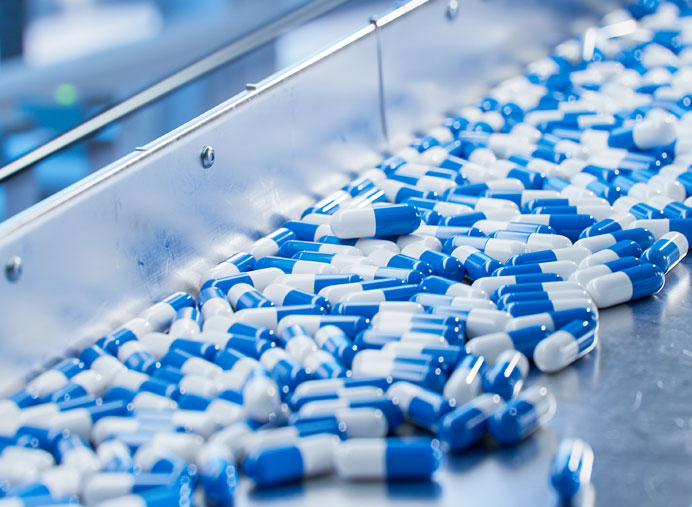 Blue and white capsules on a conveyor belt in a pharmaceutical manufacturing facility, illustrating the process of medication production.