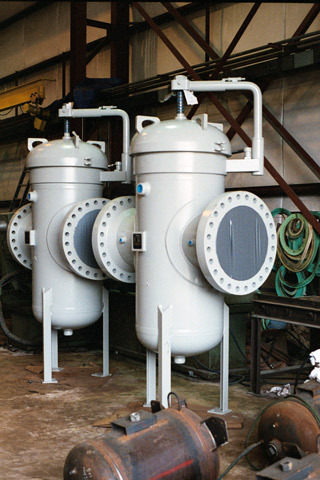 Pipeline basket strainers in an industrial setting, featuring two large gray filter units designed for fluid filtration. The strainers are mounted on sturdy legs with visible inlet and outlet connections, surrounded by hoses and equipment in a workshop environment.