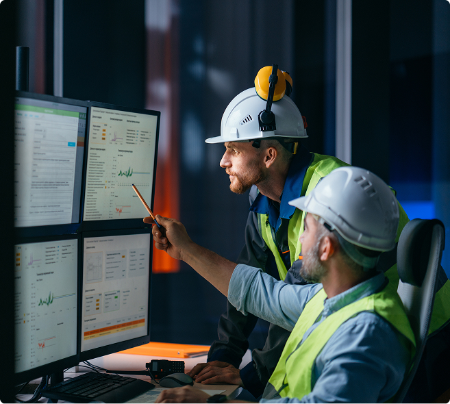 Two engineers in hard hats and safety vests analyze data on multiple computer screens in a control room, discussing findings and pointing at graphs and charts.