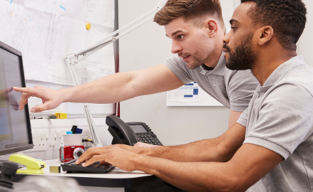 Two colleagues collaborating at a desk, one pointing at a computer screen while the other types, surrounded by office equipment and project plans.