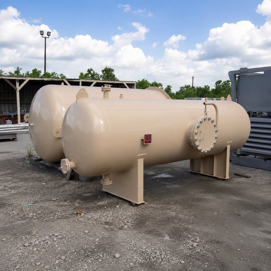 Two beige pressure vessels positioned on a gravel surface, showcasing their cylindrical design and industrial setting, with a clear blue sky and scattered clouds in the background.