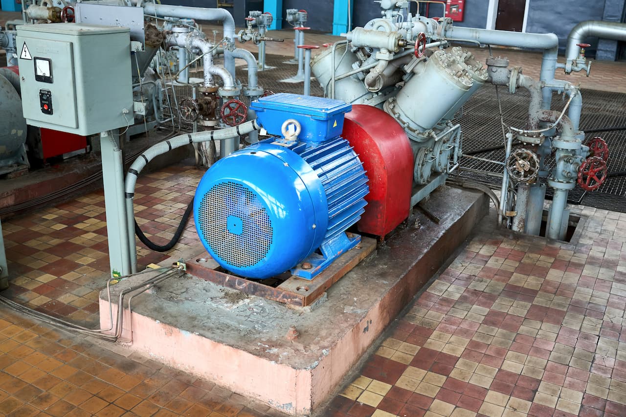 Industrial electric motor in a machinery room, featuring blue and red components, connected to a series of pipes and valves for fluid management. The floor is tiled, indicating a functional workspace for mechanical operations.
