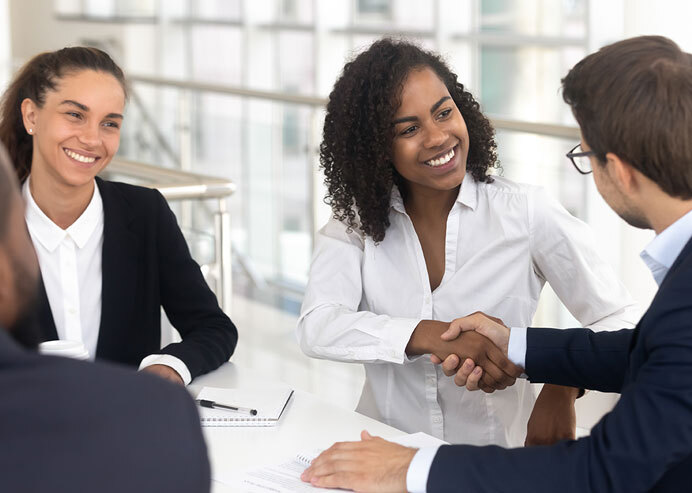 Diverse group of professionals in a meeting, discussing staffing solutions in manufacturing, with one woman smiling and shaking hands, symbolizing successful collaboration and recruitment strategies.