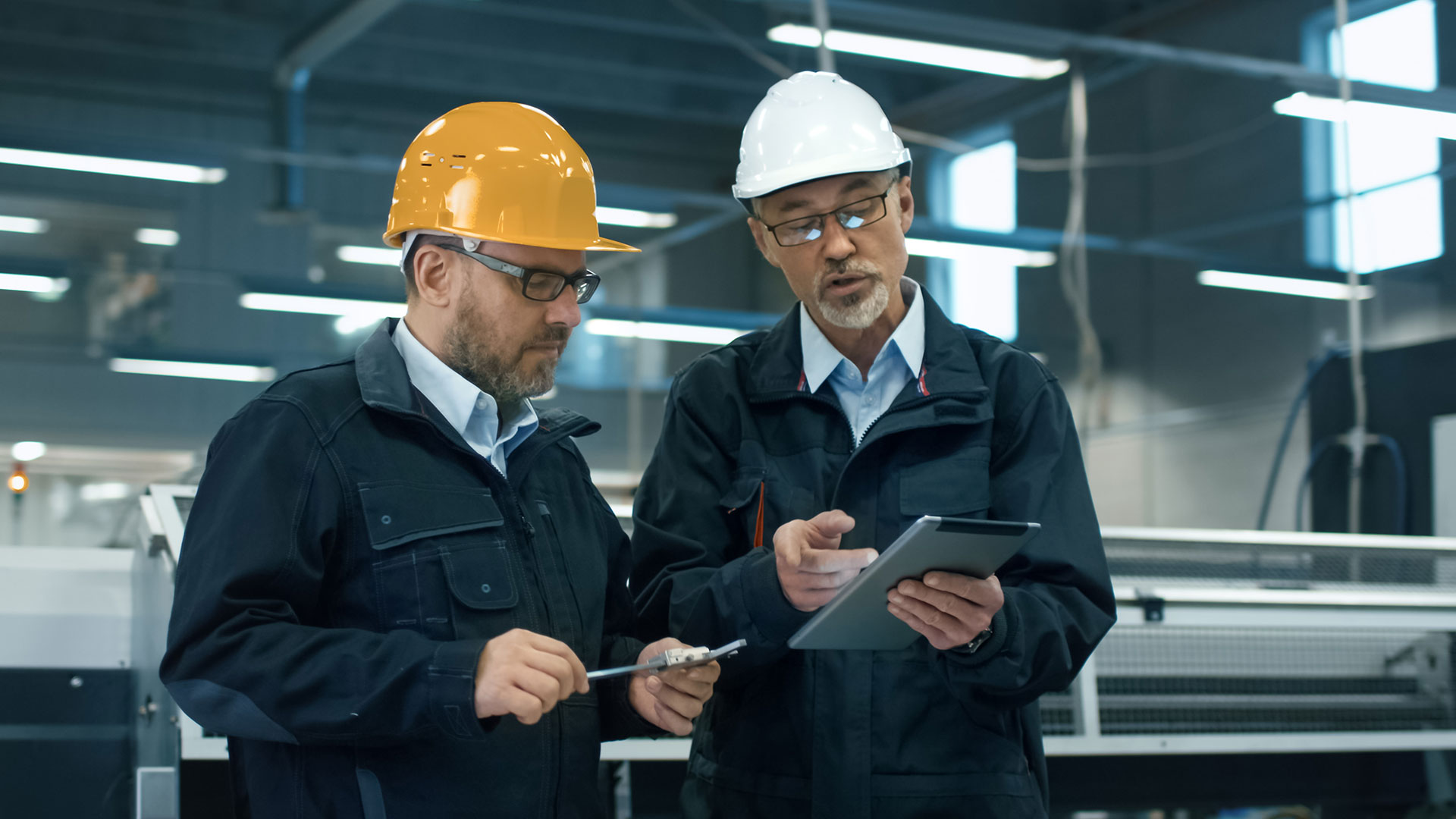 Two engineers in safety helmets discuss project details while inspecting equipment in a manufacturing facility. One holds a tablet, and the other uses a caliper, highlighting collaboration and attention to detail in industrial operations.
