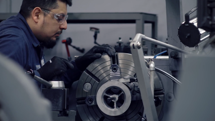 A technician wearing safety glasses and gloves operates a precision machine in a workshop, focusing on adjusting the equipment for metalworking.