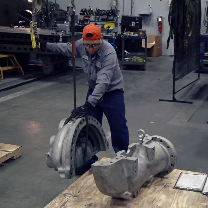 Worker in a manufacturing facility using a hoist to lift a large metal component, with additional machinery and tools visible in the background.