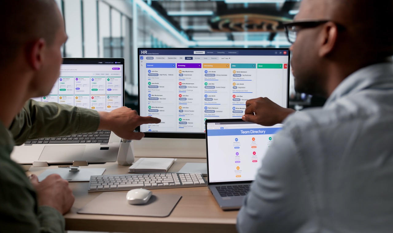Two professionals collaborating at a modern office desk, analyzing HR software displayed on multiple screens. One individual points at a candidate list on a desktop monitor while the other reviews the team directory on a laptop, highlighting teamwork and technology in recruitment processes.