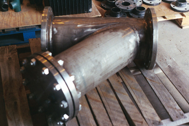 Y strainer made of stainless steel, showcasing flanged connections, resting on wooden pallets with various industrial components in the background. Ideal for filtering debris from liquids in piping systems.